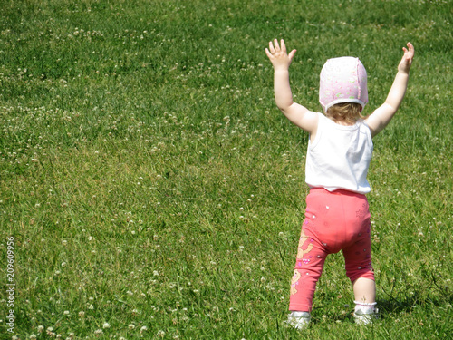 A joyful little child enjoys the summer playing in a green meadow, rear view. Little girl raised his hands up, happy childhood, baby first steps