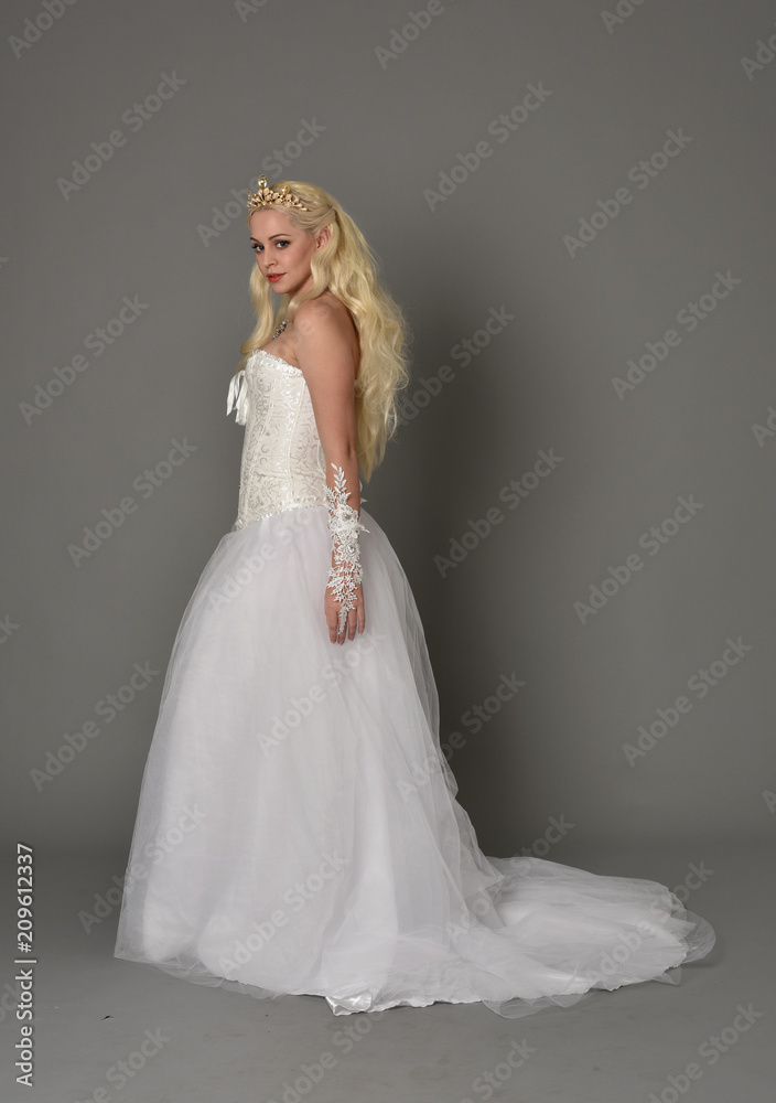 full length portrait of blonde  girl wearing white fantasy gown. standing pose in side profile, grey studio background.