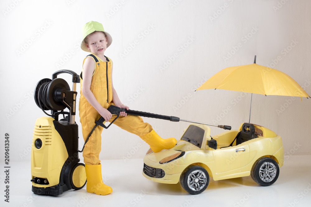 Happy child in yellow overalls and boots with high pressure washer serves client boy on toy car under umbrella. Concept of family business. White background Happy child in yellow overalls and boots with high pressure washer serves client boy on toy car under umbrella. Concept of family business. White background