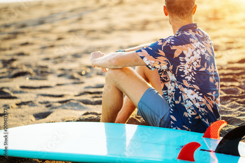 Portrait of happy surfer in hawaiian t-shirt walking with surf board on the beach at sunset