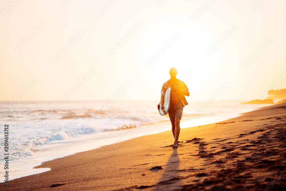 Obraz premium Portrait of happy surfer in hawaiian t-shirt walking with surf board on the beach at sunset