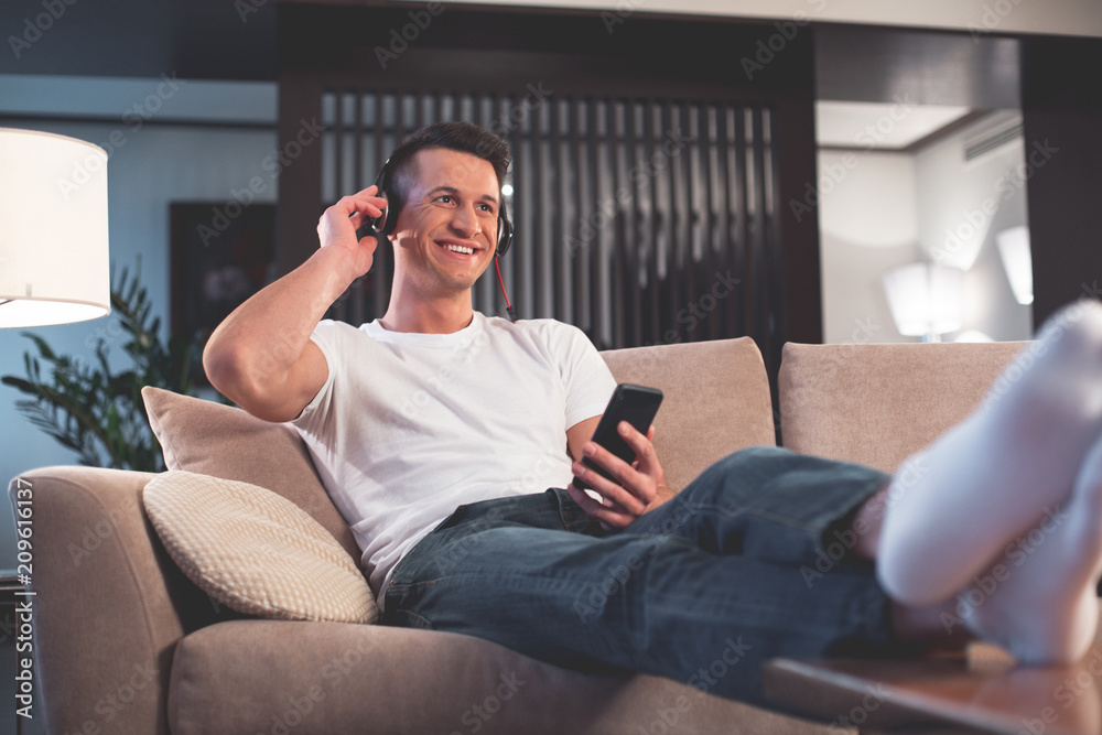 Low angle of positive guy enjoying favorite song while relaxing at home. He is sitting on couch and leaning feet on table. Male is touching headphone and laughing 