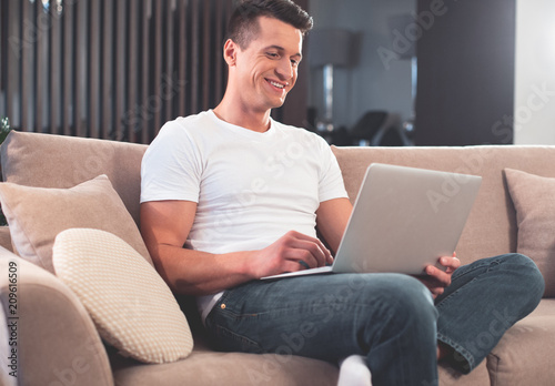 Cheerful guy is spending free time at home. He is watching video on laptop and laughing while sitting on couch 
