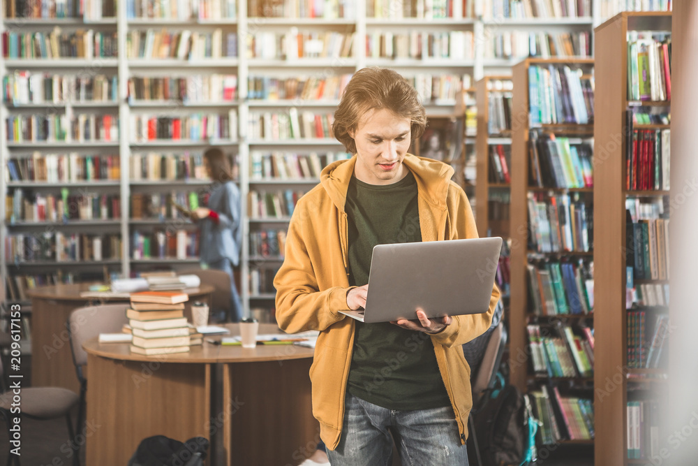 Portrait of cheerful man watching at notebook computer while standing in library near bookshelves Portrait of cheerful man watching at notebook computer while standing in library near bookshelves