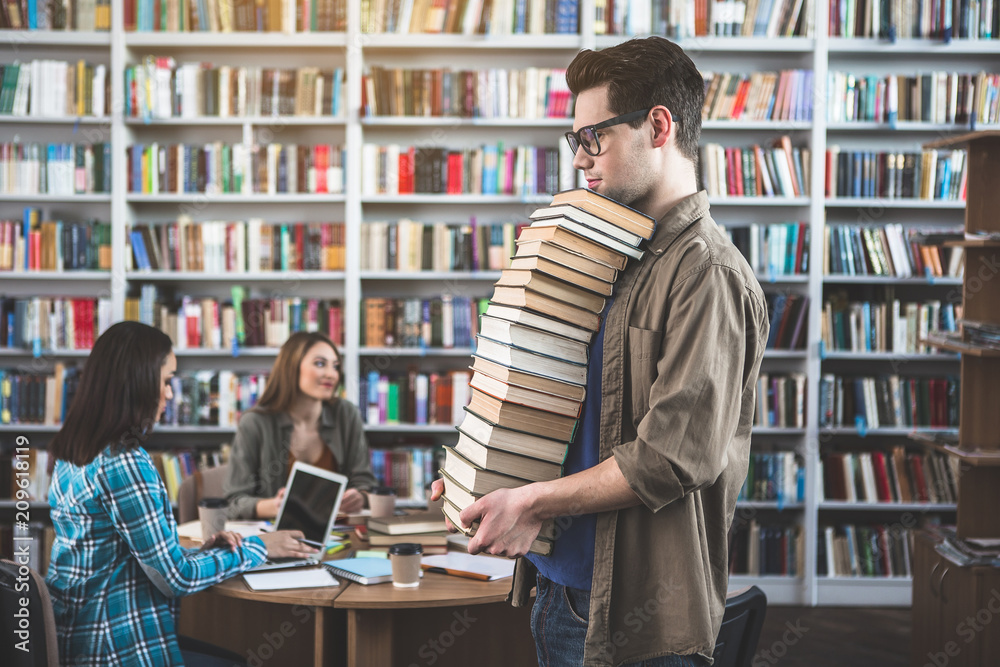 Glad male holding different volumes in hands in library. He situating near happy girls. They working with laptop and tasting cup of coffee Glad male holding different volumes in hands in library. He situating near happy girls. They working with laptop and tasting cup of coffee