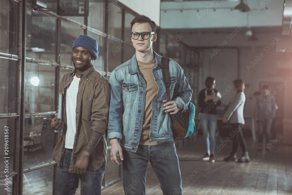 Best friends. Portrait of young cheerful stylish men with backpacks are standing in office and looking ahead confidently. Their colleagues are standing in background. Selective focus