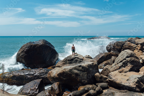 a man stands on a rock and looks at the ocean, waves of waves fly upward