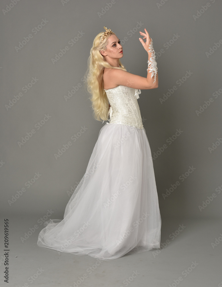 Fototapeta premium full length portrait of blonde girl wearing white fantasy gown. standing pose in side profile, grey studio background.