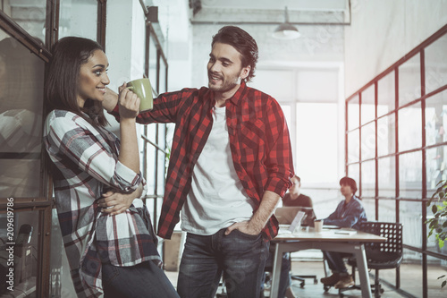Side view profile of optimistic girl is drinking espresso while leaning on glass wall. She is looking at man who is standing near and flirting with her. Professional team is laboring in background
