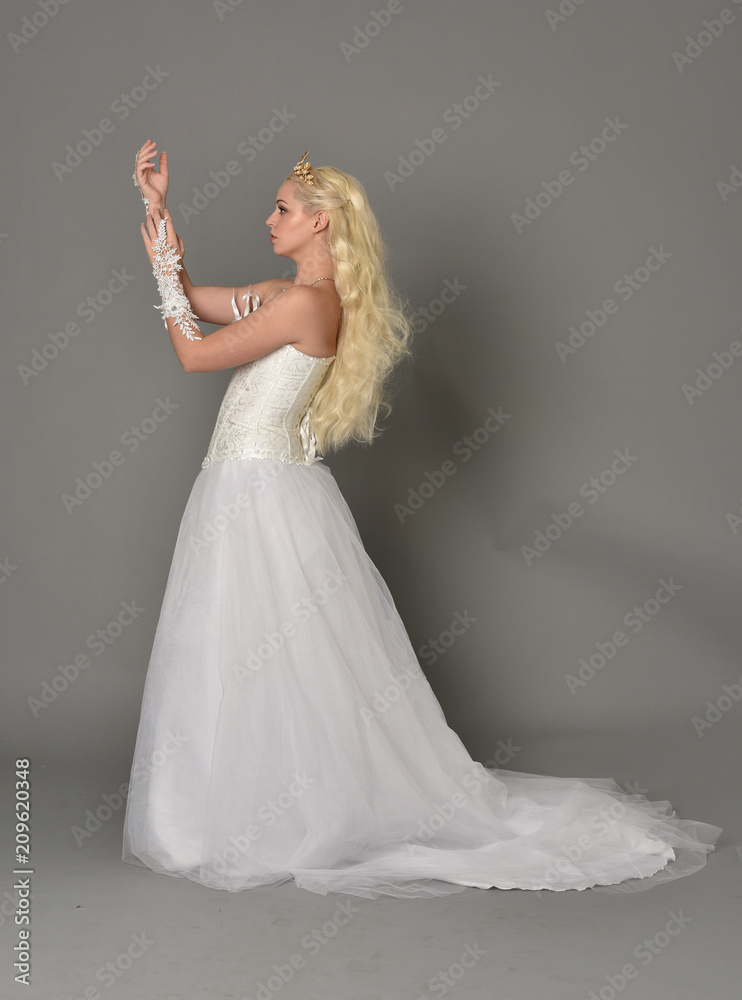 Naklejka premium full length portrait of blonde girl wearing white fantasy gown. standing pose in side profile, grey studio background.
