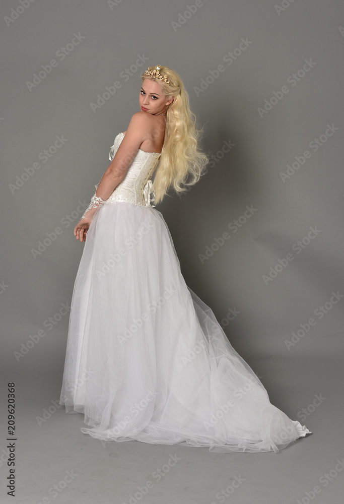 full length portrait of blonde  girl wearing white fantasy gown. standing pose in side profile, grey studio background.