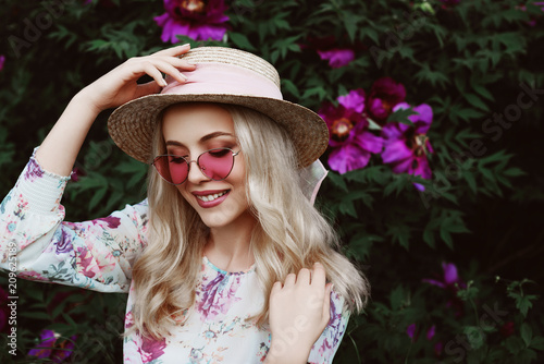 Outdoor close up portrait of beautiful young happy smiling woman with long blonde hair, wearing pink sunglasses, straw hat, dress posing in the blooming garden.  Female spring fashion concept