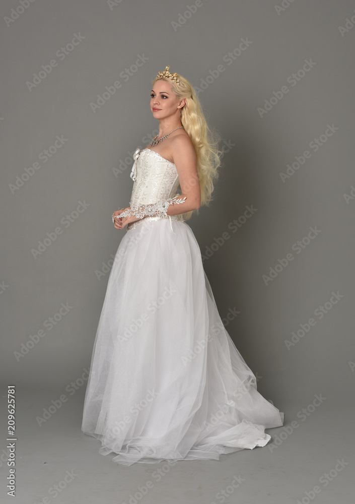 full length portrait of blonde  girl wearing white fantasy gown. standing pose in side profile, grey studio background.