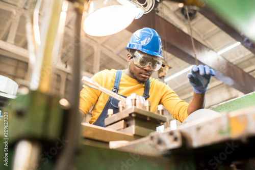 Serious busy young black factory engineer in hardhat and safety goggles examining milling lathe and repairing it while working at production plant photo