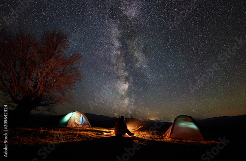 Night camping in mountains. Silhouette of male hiker sitting between two illuminated tents  enjoying campfire  beautiful starry sky and Milky way on background. Tourism  outdoor activity concept