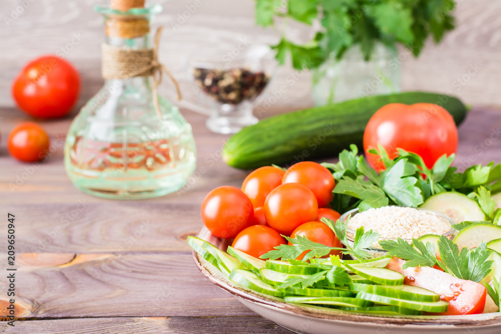 Fresh slices of cucumbers, tomatoes, sesame seeds in a bowl and parsley leaves on a plate on a wooden table. Oil and seasoning for cooking vegetable salad Fresh slices of cucumbers, tomatoes, sesame seeds in a bowl and parsley leaves on a plate on a wooden table. Oil and seasoning for cooking vegetable salad