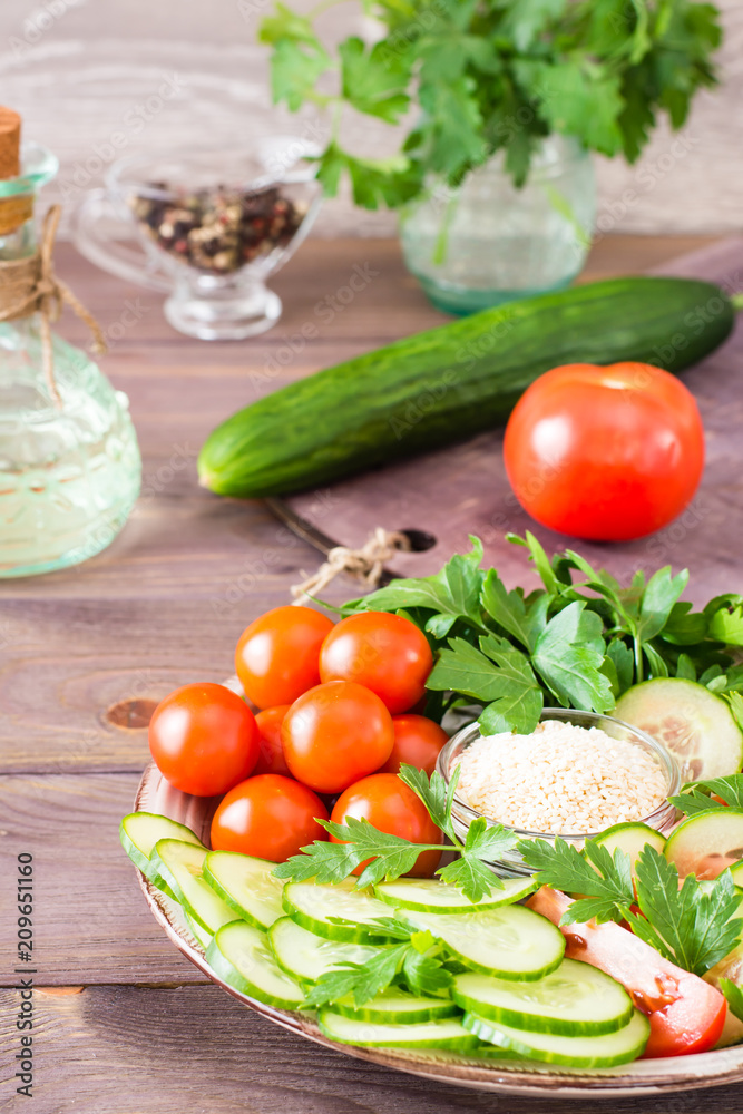 Fresh slices of cucumbers, tomatoes, sesame seeds in a bowl and parsley leaves on a plate on a wooden table. Oil and seasoning for cooking vegetable salad Fresh slices of cucumbers, tomatoes, sesame seeds in a bowl and parsley leaves on a plate on a wooden table. Oil and seasoning for cooking vegetable salad