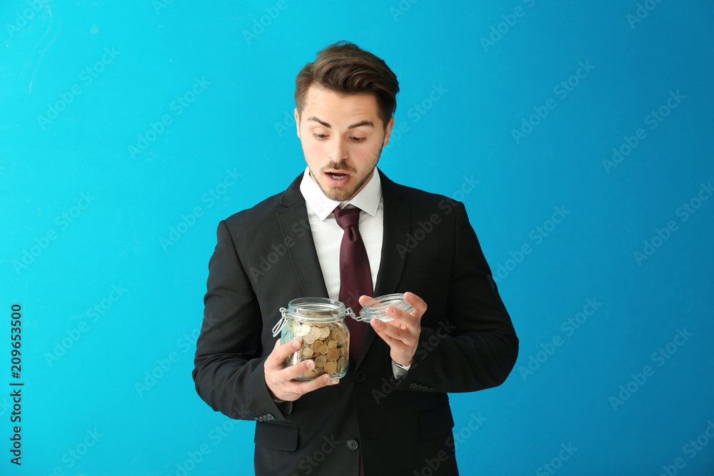 Young surprised businessman holding glass jar with coins on color background. Savings money concept Young surprised businessman holding glass jar with coins on color background. Savings money concept