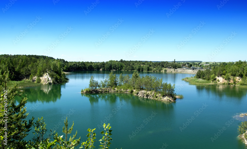 An island in the shape of an arc in the middle of a large lake near the rocks, Blue clear sky An island with trees and forestland, Reflection in the water