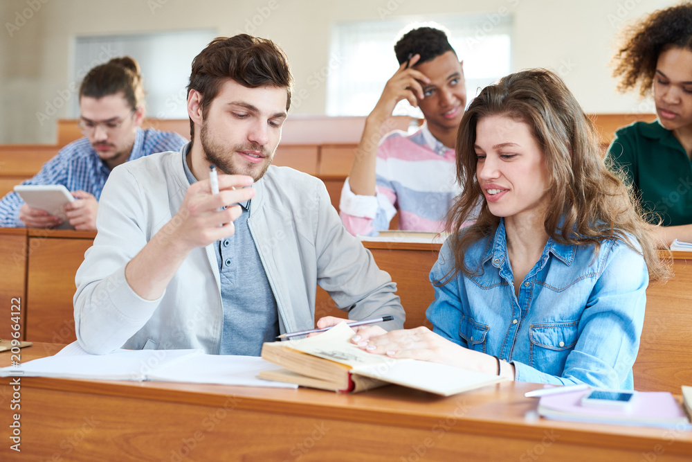Cheerful positive student friends analyzing textbook and discussing main idea while sharing thought during break in lecture room Cheerful positive student friends analyzing textbook and discussing main idea while sharing thought during break in lecture room