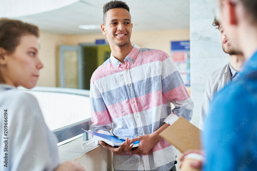 Cheerful positive handsome Hispanic guy in stripped shirt holding workbook and leaning on railing while talking to university friends in lobby Cheerful positive handsome Hispanic guy in stripped shirt holding workbook and leaning on railing while talking to university friends in lobby