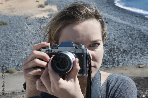 Young millennial female of Caucasian ethnicity holding a digital, professional camera over her right eye and taking a photograph facing forward, against a pebble beach background in a sunny day © Ana