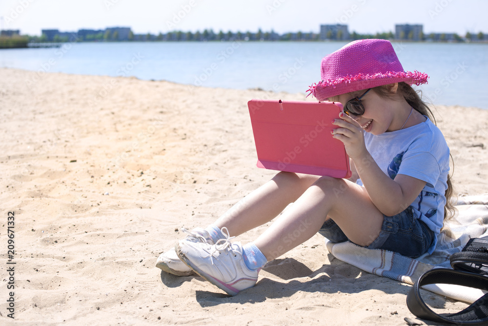 Little girl with a tablet computer in hands. Sunny day on the beach and blue sky. Girl in a pink hat and glasses. Copy space