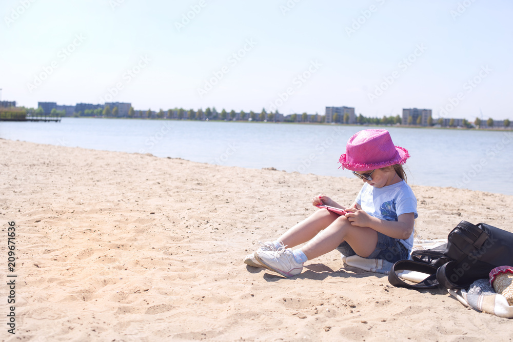 Little girl is sitting on the beach with a tablet computer in hands. Sunny day and blue sky. Girl in a pink hat and glasses. Little girl is sitting on the beach with a tablet computer in hands. Sunny day and blue sky. Girl in a pink hat and glasses.