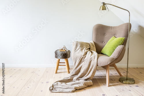 A reading chair / armchair with a lamp, blanket and a open book. Empty white wall in simple living room interior. Copy space