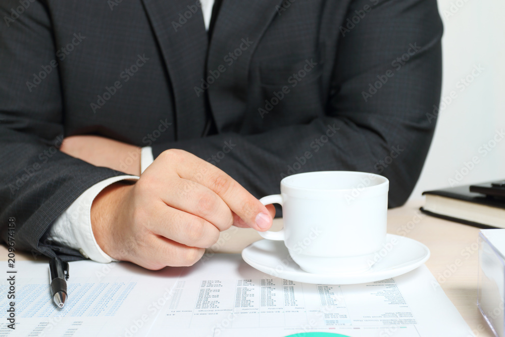 Hands of businessman with cup of coffee at table with reports, documents Hands of businessman with cup of coffee at table with reports, documents