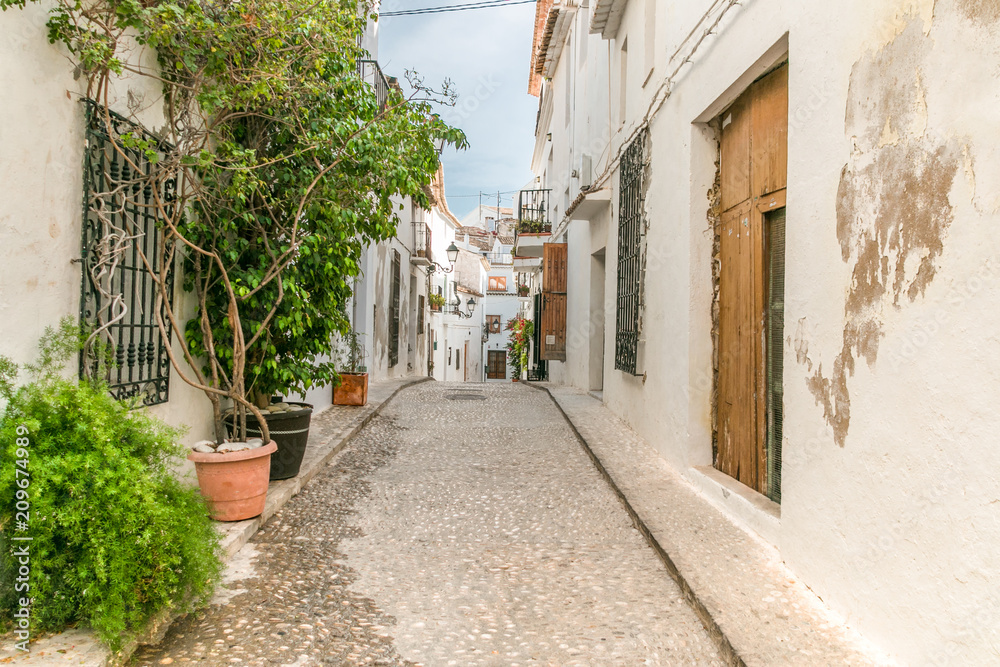A narrow, quiet stone street in the city of Altea in Valencia, Spain, Costa Blanca. On the sides houses with bright facades and plants in pots.