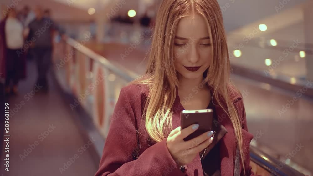 Pretty girl in coat taking escalator, looking at screen of cellphone. Young stylish woman with cherry lipstick using smartphone, browsing. Indoors. Pretty girl in coat taking escalator, looking at screen of cellphone. Young stylish woman with cherry lipstick using smartphone, browsing. Indoors.