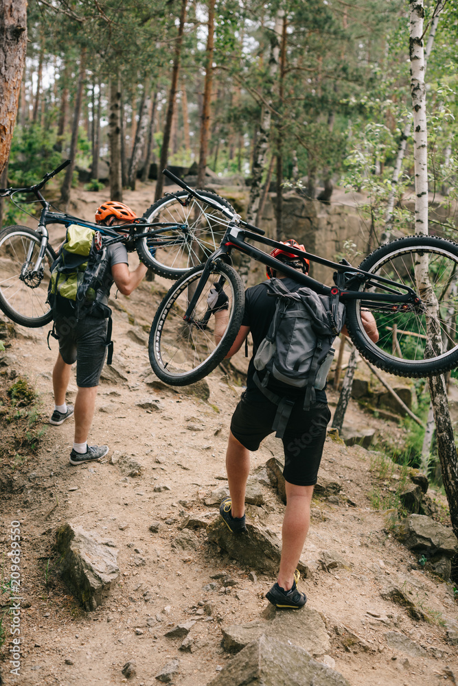 Naklejka premium rear view of trial bikers carrying bikes on backs at forest and going uphill