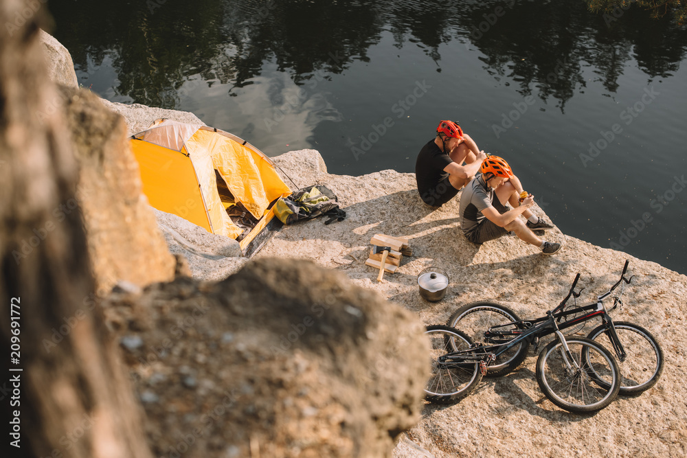 Fototapeta premium high angle view of active trial bikers eating canned food in camping on rocky cliff