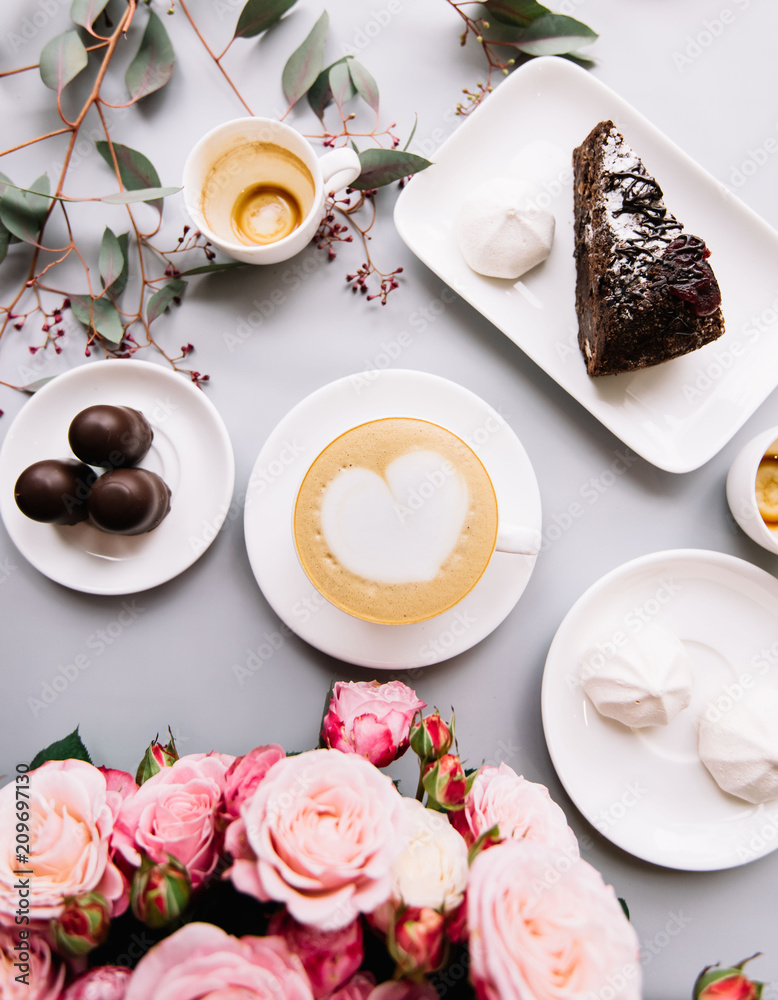 Delicious fresh morning cappuccino coffee with heart shaped latte art on it, a piece chocolate cake, marshmallow, chocolate, empty cups, eucalyptus and a big bouquet of pink roses, flat lay, top view.