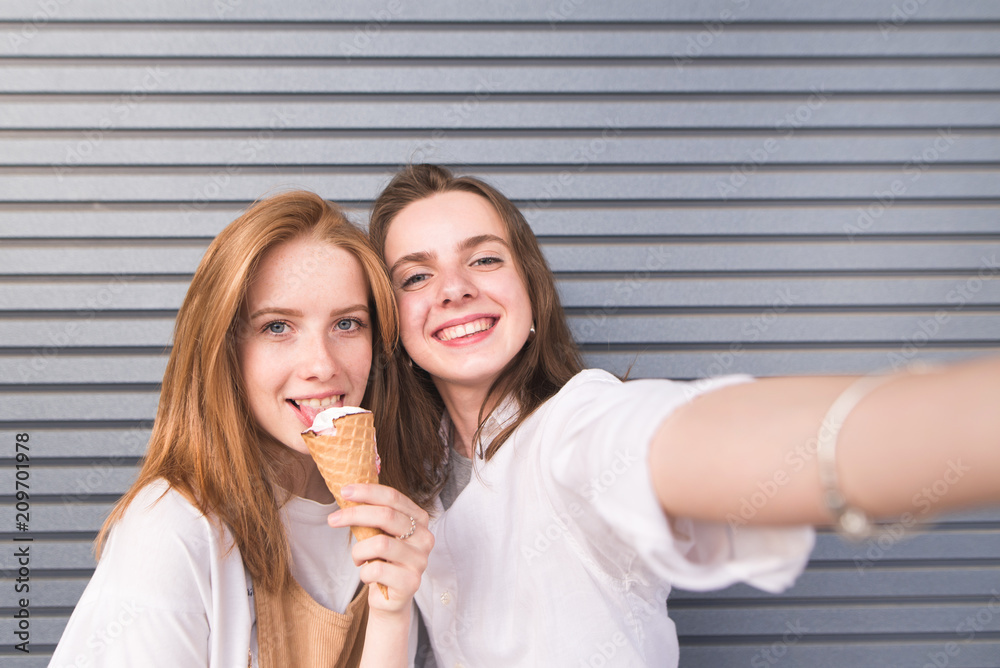 Cute happy young women in bright clothes make selfie on a dark background. Attractive young girl doing selfie with ice cream on a background of gray walls. Girlfriends are photographed on a smartphone Cute happy young women in bright clothes make selfie on a dark background. Attractive young girl doing selfie with ice cream on a background of gray walls. Girlfriends are photographed on a smartphone
