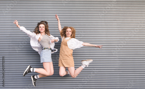 Happy young girls jumping with their hands raised on a gray dark background. Two smiling girlfriends isolated in a jump against the backdrop of a gray wall. Copyspace