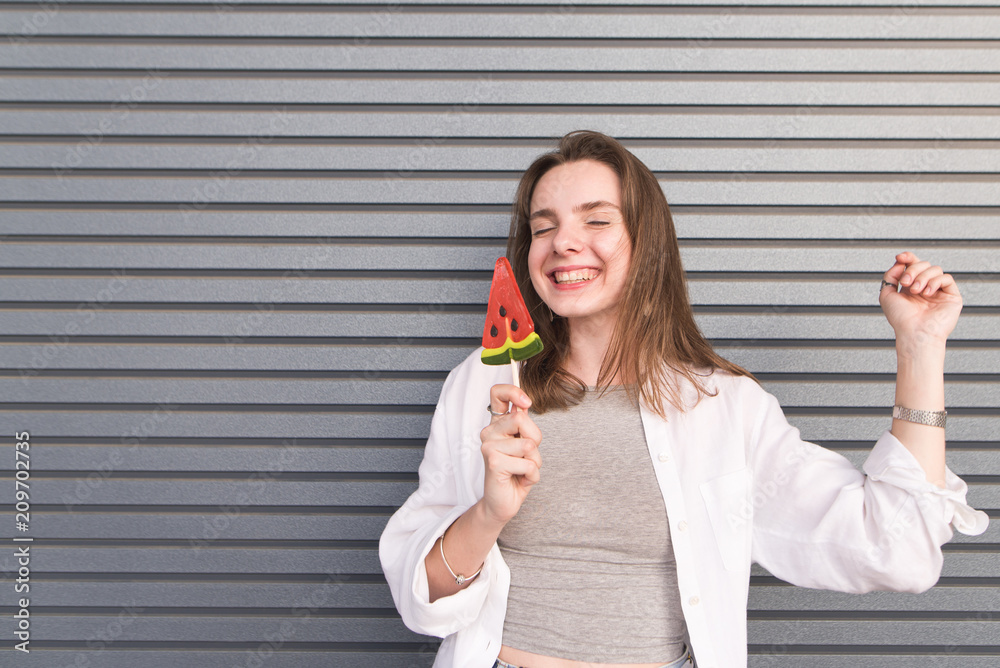 Happy girl standing with a lollipop in her hand and smiling warmly on a gray background. Portrait of smiling happy girl with candy on the background of the wall Happy girl standing with a lollipop in her hand and smiling warmly on a gray background. Portrait of smiling happy girl with candy on the background of the wall