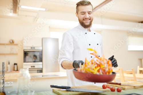 Portrait of modern professional chef mixing ingredients in frying pan while working  in modern restaurant kitchen, copy space