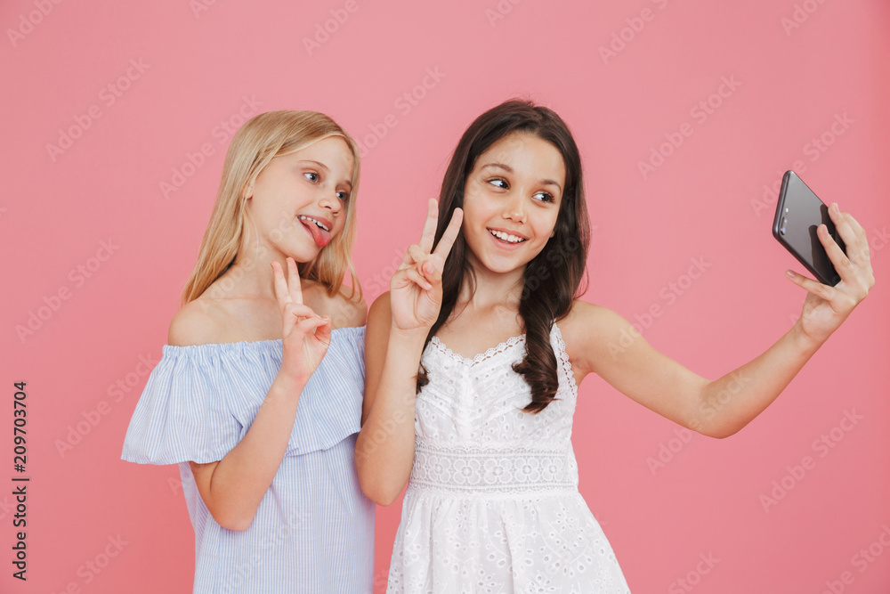 Picture of brunette and blonde adorable girls wearing dresses smiling and showing peace sign while taking selfie on mobile phone, isolated over pink background Picture of brunette and blonde adorable girls wearing dresses smiling and showing peace sign while taking selfie on mobile phone, isolated over pink background