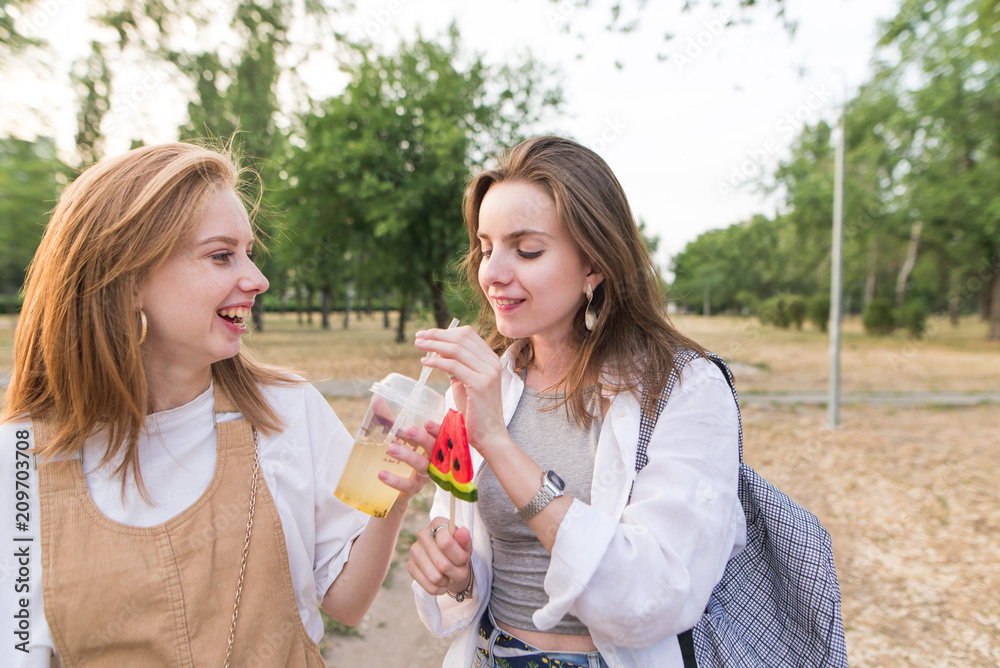 Two positive girls with a lemonade and a lollipop are walking around the park. Portrait of stylish happy girls in the park. Walk with a girlfriend. Two positive girls with a lemonade and a lollipop are walking around the park. Portrait of stylish happy girls in the park. Walk with a girlfriend.