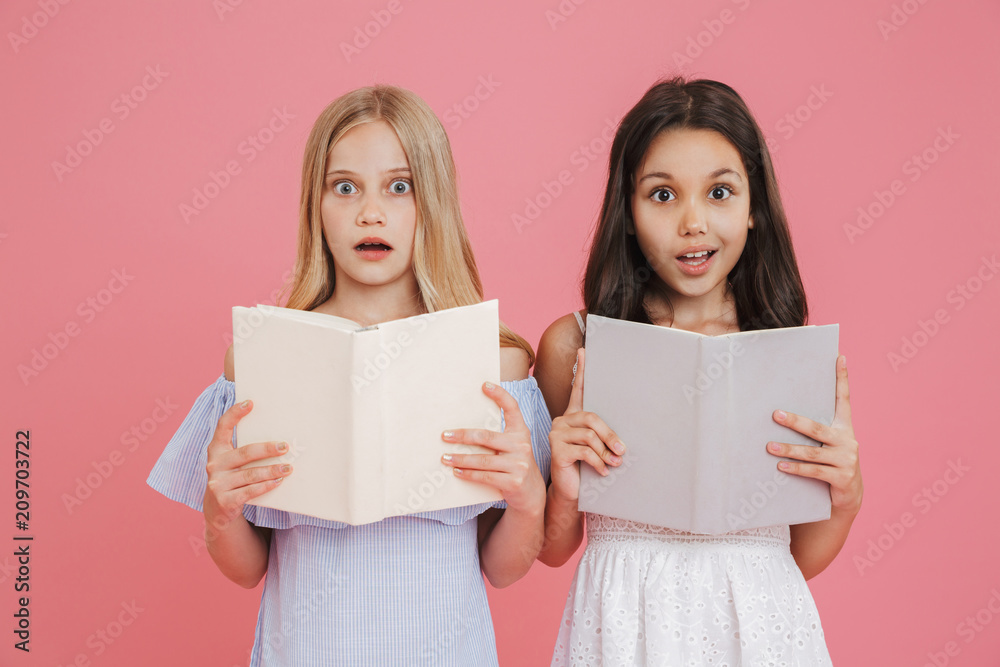 Photo of brunette and blonde schoolgirl wearing dresses holding and reading books together with excitement, isolated over pink background Photo of brunette and blonde schoolgirl wearing dresses holding and reading books together with excitement, isolated over pink background