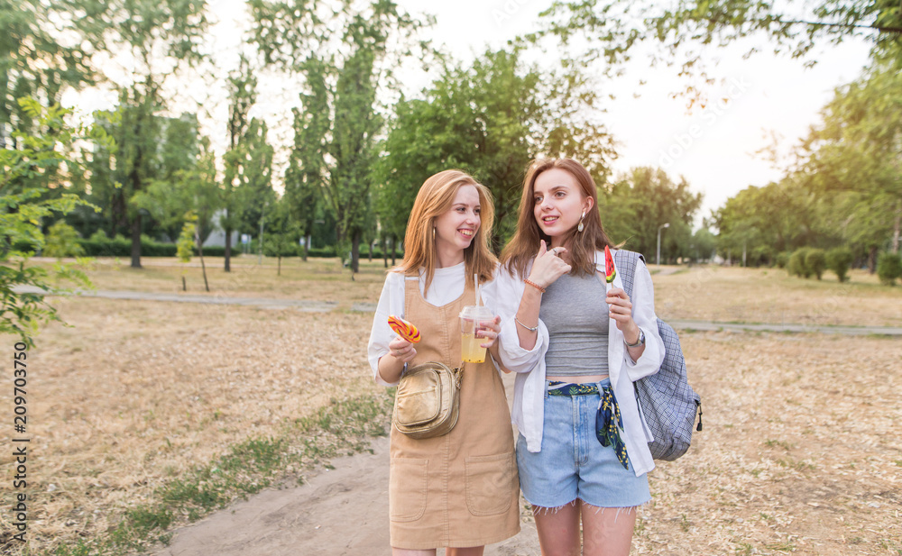 Fototapeta premium Two stylish girlfriends in light clothing with lemonade and lollipops stroll through the summer evening park. Two stylish attractive girls walking around the park and smiling.