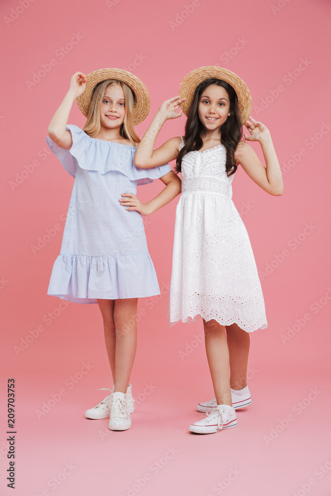 Full length portrait of beautiful summer girls 8-10 wearing dresses posing at camera touching straw hats with smile, isolated over pink background Full length portrait of beautiful summer girls 8-10 wearing dresses posing at camera touching straw hats with smile, isolated over pink background