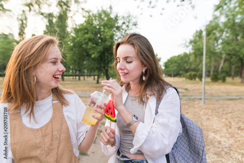 Two positive girls with a lemonade and a lollipop are walking around the park. Portrait of stylish happy girls in the park. Walk with a girlfriend.