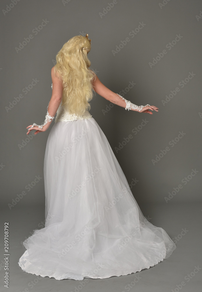 full length portrait of blonde girl wearing white gown, standing pose with back to the camera. grey studio background. full length portrait of blonde girl wearing white gown, standing pose with back to the camera. grey studio background.