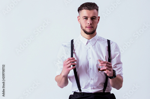 Confident, stylish man in white shirt and suspenders looking to camera and holding glass of whiskey in hand while sitting against gray background. Elegant man drinking alcohol in studio. Men's fashion