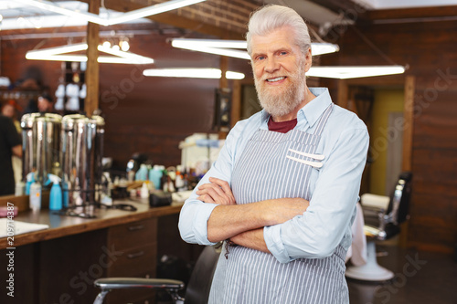 Stylish uniform. Delighted gray-haired barber expressing positivity while standing at his workplace