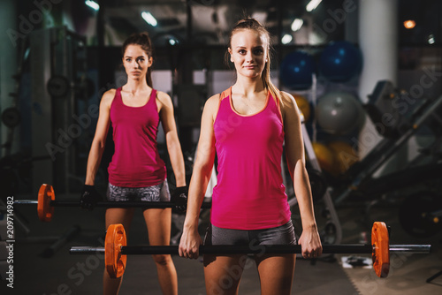 Two gorgeous girls in pink tank tops are holding weights in a gym with dimmed light.