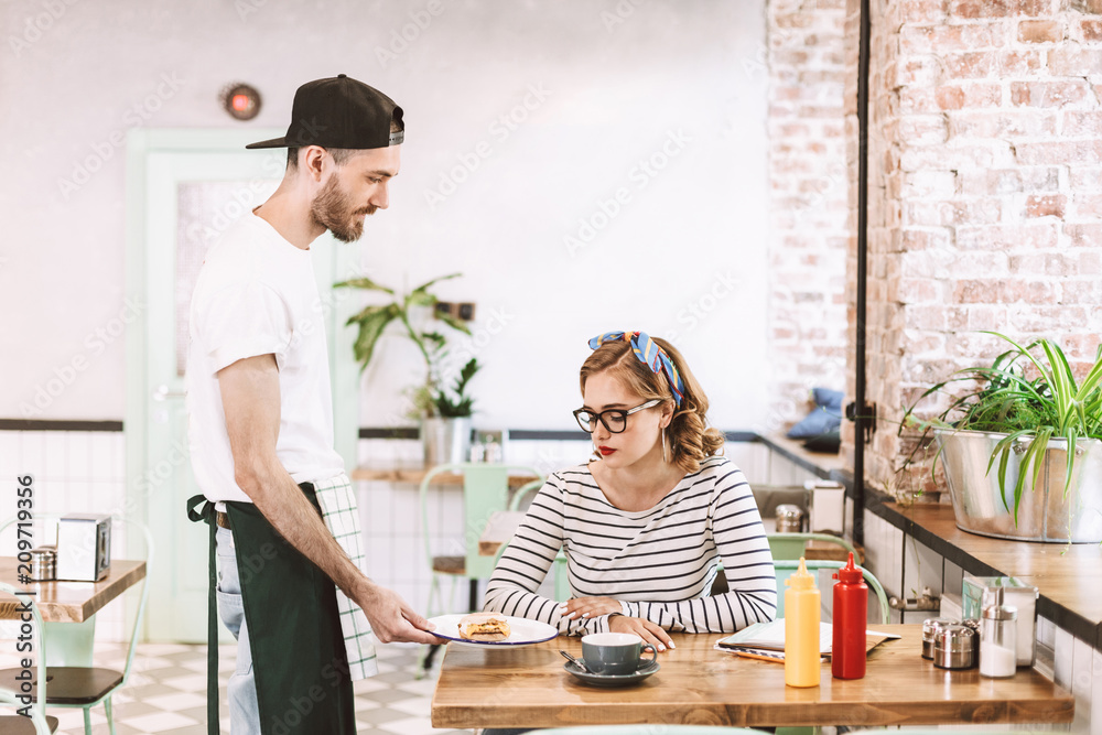 Waiter in black cap giving burger to pretty lady in eyeglasses which sitting at the table in cafe